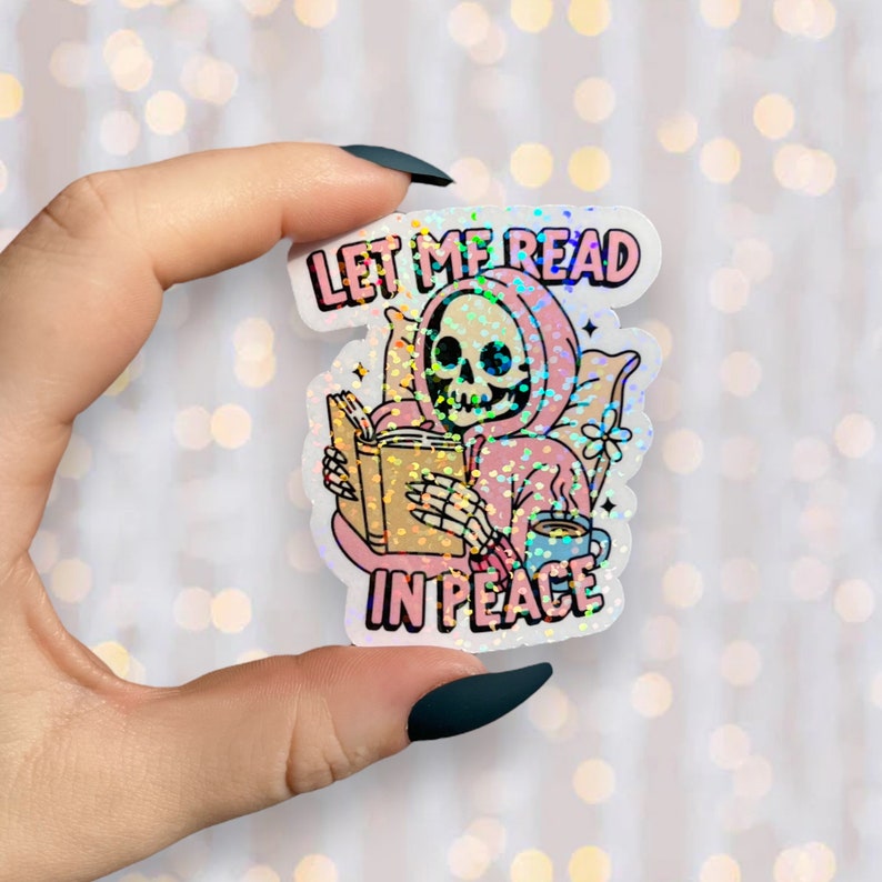May include: A holographic sticker showing a skeleton in a pink hoodie reading a book and drinking coffee. The sticker reads "LET ME READ IN PEACE" in pink and white text. The sticker is held by a hand with dark green nail polish.