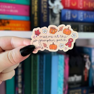 May include: A sticker with a fall theme, featuring pumpkins, leaves, a mug, and a scarf. The text "meet me at the pumpkin patch" is written on the sticker. The sticker is held by a person with black nail polish.