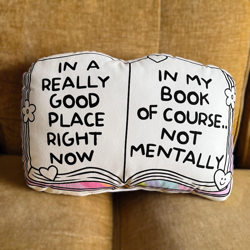 May include: White book-shaped pillow with black text that reads "In a really good place right now" on the left side and "In my book of course... not mentally" on the right side. The pillow has a rainbow design on the inside of the book.