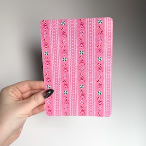 May include: A pink rectangular card with a repeating pattern of red bows, green and white peppermint candies, and white starbursts. The card has rounded corners and is held by a hand with a black and silver nail design.