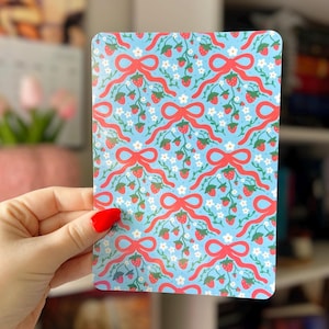 May include: A rectangular card with a light blue background featuring a repeating pattern of red bows, strawberries, and white flowers. The card is held by a hand with red nail polish.