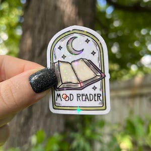 May include: A white sticker with a holographic finish features a black and white illustration of an open book with a crescent moon above it. The text "Mood Reader" is printed below the book.
