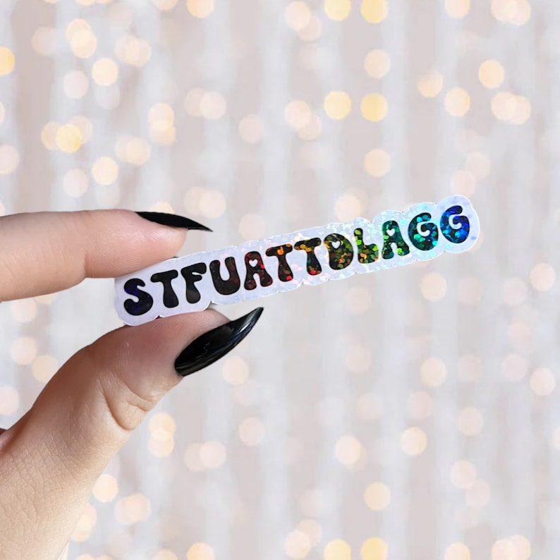 May include: A white sticker with the text "STFUATTLAGG" in black, rainbow glitter letters. The sticker is being held by a hand with black nail polish.