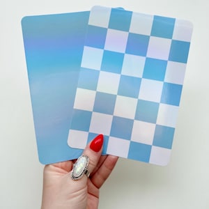 May include: Two rectangular decorative boards in shades of blue. One has a gradient effect, the other a checkered pattern of light and dark blue squares. The boards are held by a hand with a red nail and a silver ring. The boards are likely for home decor.