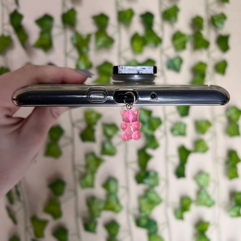 May include: A pink gummy bear charm attached to a black phone case with a pop socket.