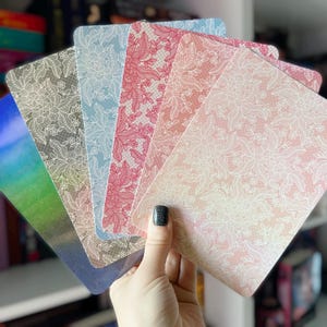 May include: A set of six decorative cards featuring a lace pattern. The cards are in various shades of pink, blue, and white.