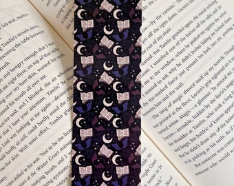 Puede incluir: Un marcapáginas con un patrón repetido de libros abiertos, lunas crecientes y murciélagos sobre un fondo azul oscuro. Los libros son morados y blancos, las lunas son blancas y los murciélagos son morados.