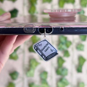 Puede incluir: Una funda transparente para teléfono con un dije de libro negro que dice "Drarry Fanfic" con dos corazones sobre el título. El dije está unido a la funda del teléfono con un anillo plateado.