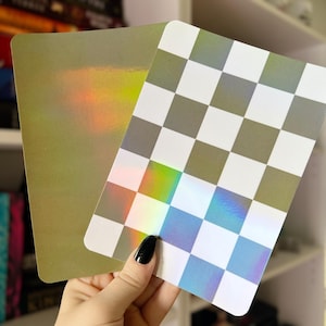 May include: Two rectangular cards with iridescent surfaces. One card has a gold finish, while the other features a checkerboard pattern in white and gray. The cards reflect a rainbow of colors, creating a holographic effect.