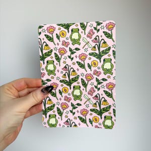 May include: A rectangular pink surface with a repeating pattern of green frogs, pink and yellow flowers, butterflies, dragonflies, and stars. The design is whimsical and colorful, with a cartoonish aesthetic.