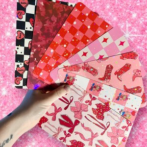 Puede incluir: Un conjunto de seis hojas de papel con patrones de purpurina rosa y roja. Los patrones incluyen corazones, estrellas, cuadros y botas de vaquero.