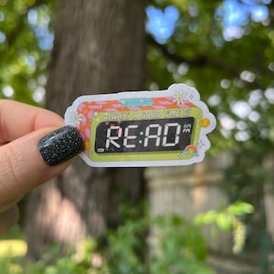 May include: A colorful sticker with a digital clock that reads "RE:AD" and the text "Always a good time to". The clock has a pink and white checkered background and a yellow face with a rainbow and stars.