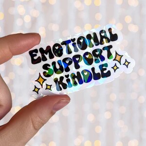 May include: A holographic sticker with the text "Emotional Support Kindle" in black lettering with a rainbow iridescent background. The sticker has two small yellow stars on the bottom right corner.