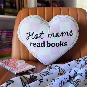 May include: A pink and white heart-shaped pillow with the text "Hot moms read books" on it. The pillow is sitting on a brown leather chair.