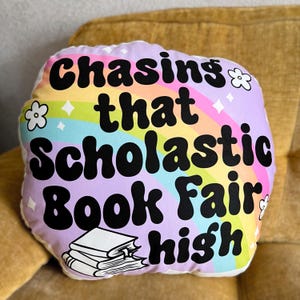 May include: A white pillow with a rainbow and stars design. The text "Chasing that Scholastic Book Fair high" is printed in black on the pillow.