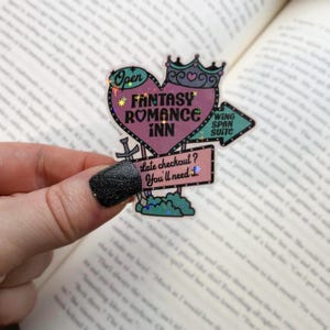 Könnte beinhalten: Ein rosafarbenes herzförmiges Schild mit dem Text "Fantasy Romance Inn" und einer Krone oben drauf. Das Schild ist von einem grünen Rand umgeben und hat ein kleineres Schild darunter, auf dem steht "Late checkout? You'll need a..."