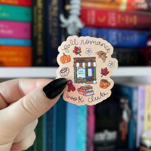 May include: A holographic sticker with a fall theme. The sticker features a pumpkin, maple leaves, a coffee cup, and a bookstore cafe. The text "fall romance" and "book club" are also included in the design.
