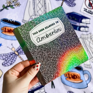 May include: A black and iridescent notebook with a rainbow effect. The notebook has a white label with the text "This Kobo belongs to Ambertin".