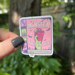 May include: A pink and green sticker of a book with the title "Monster Romance". The book cover features a green hand holding a red heart.
