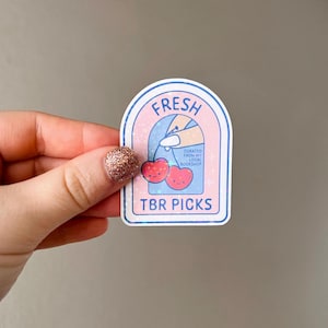 May include: A pink and blue sticker with a white border. The sticker has a white arch with the text "FRESH" in blue letters. Inside the arch is a hand holding a cherry with a smiling face. The text "CURATED FROM MY LOCAL BOOKSHOP" is in black letters. Below the cherry is the text "TBR PICKS" in blue letters.
