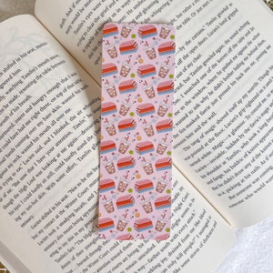 Könnte beinhalten: Rosafarbenes Lesezeichen mit einem sich wiederholenden Muster aus gestapelten Büchern und Tassen Bubble Tea. Das Lesezeichen hat einen weißen Rand und den Text "The Night Court, My Delight" unten.