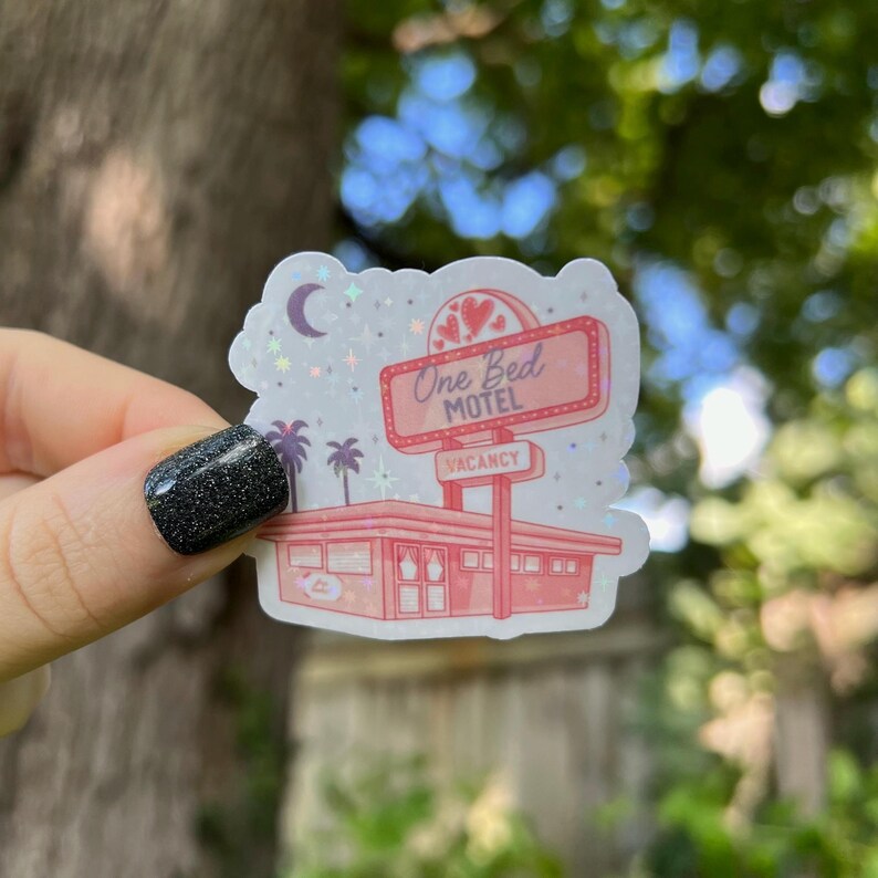 May include: A pink and white sticker with a retro motel sign that reads "One Bed Motel" with a vacancy sign. The sticker has a glittery finish and features a crescent moon and stars.