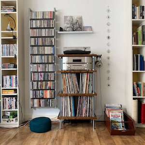 May include: A home entertainment setup featuring a turntable, amplifier, and speakers. The room includes multiple bookshelves filled with CDs, DVDs, and books. A small, teal-colored ottoman sits on the wooden floor.