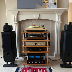 May include: A home audio system featuring a wooden shelf unit with a turntable, amplifier, and other components. Two large black speakers flank the unit. A colorful rug sits on the floor in front of the system. A painting hangs above the fireplace.