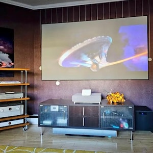 May include: A home entertainment setup featuring a large screen displaying a spaceship image. A dark wood cabinet with glass doors holds electronics, and a subwoofer is on the right. A multi-tiered shelf unit is on the left.