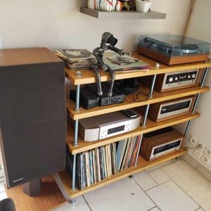 May include: A wooden audio rack with multiple shelves, holding a turntable, stereo components, and vinyl records. A large speaker is on the left. A metal sculpture sits on top of the rack. The rack is made of light-colored wood.