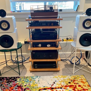 May include: A home audio setup featuring two white speakers with black and purple accents, positioned on either side of a wooden shelf unit. The unit holds a turntable and various audio components. A colorful rug and small side tables complete the scene.