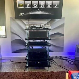 May include: A black and silver audio-visual equipment rack with multiple shelves. The rack holds various electronic components, including a TV screen, a receiver, and a DVD player. The rack is positioned in front of a wall with a decorative panel.