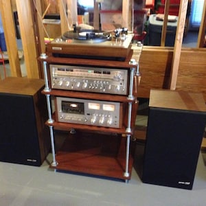 May include: A vintage stereo system featuring a wooden turntable, receiver, and cassette deck on a custom wooden and metal stand. Two black speakers with wooden tops flank the stand, creating a classic audio setup.