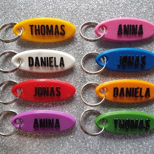 Könnte beinhalten: Acht ovale Schlüsselanhänger aus Kunststoff in verschiedenen Farben mit schwarzer Beschriftung. Die Schlüsselanhänger sind mit den Namen Thomas, Anina, Daniela und Jonas beschriftet.