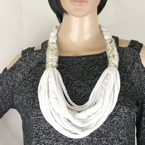 May include: A white fabric necklace with a layered, rope-like design. The necklace is worn by a person with short black hair.