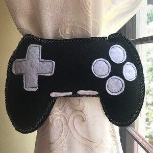 May include: Black felt video game controller curtain tieback with white buttons. The controller has a plus sign button and four circle buttons.