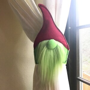 May include: A green and red gnome-shaped curtain tieback. The gnome has a red hat, a green beard, and a green pom-pom nose.