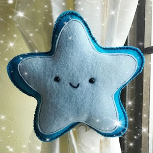 May include: A blue felt star with a smiling face, used as a curtain tieback. The star has a teal blue outline and is hanging on a white curtain.