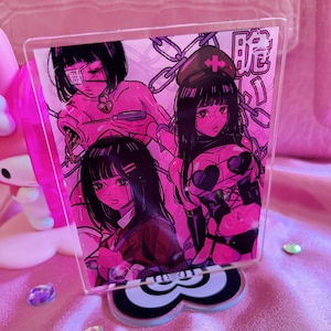 May include: A pink acrylic stand featuring a manga-style illustration of three female figures with black hair and pink accents. The stand has a black and white base with a cloud-like shape. The background is a pink satin fabric.