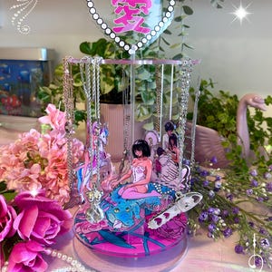 Puede incluir: Un expositor de joyas giratorio de acrílico transparente con un remate en forma de corazón que muestra el kanji japonés para el amor. La base y los paneles centrales están decorados con coloridas ilustraciones de estilo anime, un cocodrilo y motivos florales.