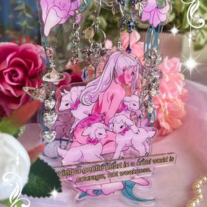 May include: A clear acrylic earring display featuring a pink illustration of a woman with long hair and lambs. The display has a floral design with the text "Having a gentle heart in a cruel world is courage, not weakness."