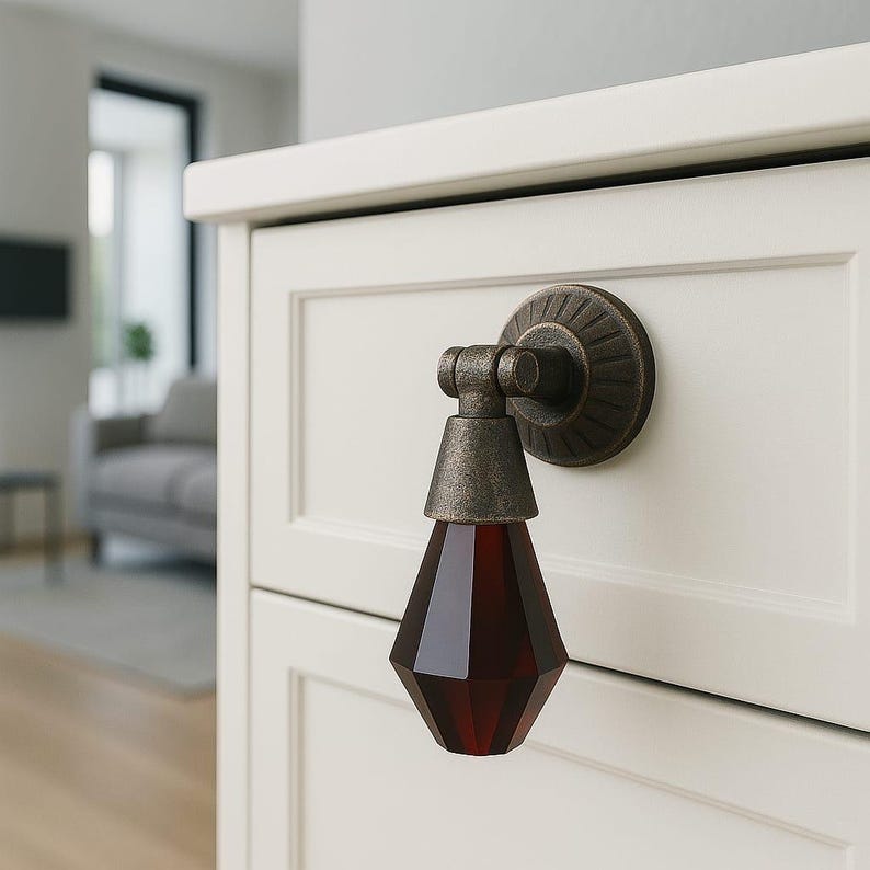 May include: A pendant-style drawer pull featuring a dark, faceted, teardrop-shaped glass element in a deep red hue. The hardware is constructed from an aged, metallic bronze-toned material with a circular, ribbed mounting plate attached.
