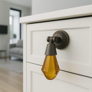 May include: A drop-style cabinet pull featuring a dark bronze-toned metal mounting plate with a circular, ribbed design and a hinged arm that holds a faceted, amber-colored glass pendant shaped like a teardrop, attached to a white drawer front.