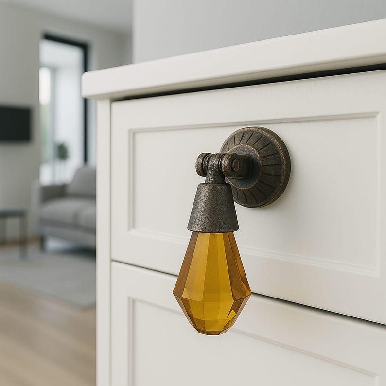 May include: A drop-style cabinet pull featuring a dark bronze-toned metal mounting plate with a circular, ribbed design and a hinged arm that holds a faceted, amber-colored glass pendant shaped like a teardrop, attached to a white drawer front.