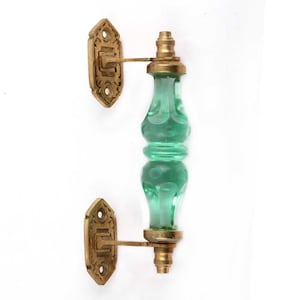 Pode incluir: Um puxador de porta com um centro de vidro verde translúcido de forma texturizada e bulbosa. O puxador é fixado por duas placas de montagem de latão retangulares e ornamentadas na parte superior e inferior, cada uma com dois orifícios.