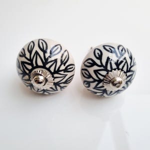 May include: Two ceramic cabinet knobs with a cream-colored base and a black floral design. Each knob has a silver-colored metal base and screw. The knobs are round and have a smooth, glossy finish.