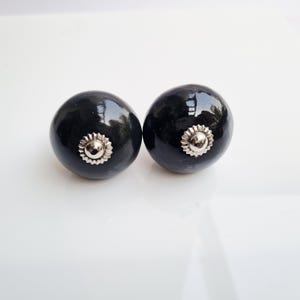 May include: Two black ceramic cabinet knobs with a shiny finish. Each knob has a silver-colored, sunburst-shaped metal accent in the center. The knobs are round and appear to be new.