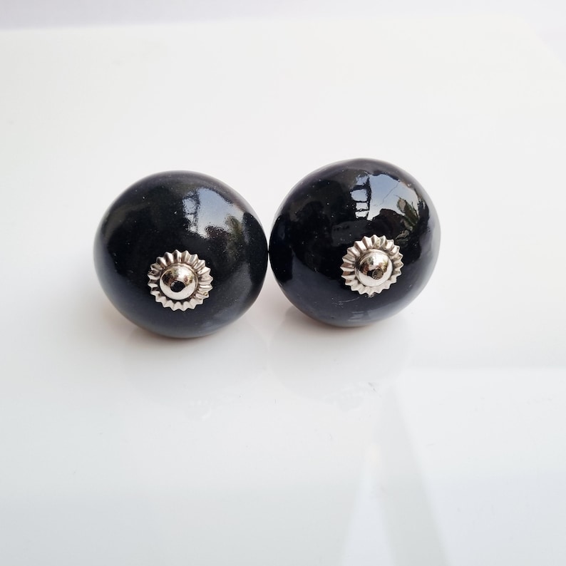May include: Two black ceramic cabinet knobs with a shiny finish. Each knob has a silver-colored, sunburst-shaped metal accent in the center. The knobs are round and appear to be new.