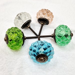 Op de afbeelding: Vijf glazen kastknoppen met een getextureerd oppervlak. De knoppen zijn in verschillende kleuren, waaronder groen, blauw, roze en helder. Elke knop heeft een messing basis en een schroef.