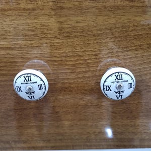 May include: Two white cabinet knobs with a clock face design. The knobs feature Roman numerals and the text "Potter-Stone". The knobs are mounted on a wooden surface.